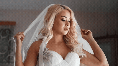 A Blonde Woman In A Wedding Dress Is Adjusting Her Veil GIF