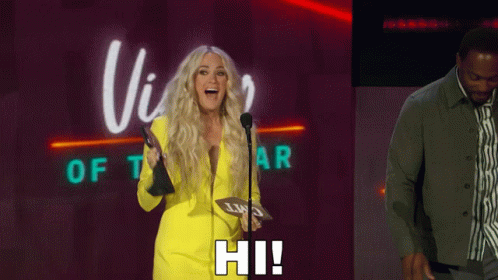 A Blonde Woman In A Yellow Suit Stands In Front Of A Microphone And Says Hi ! GIF