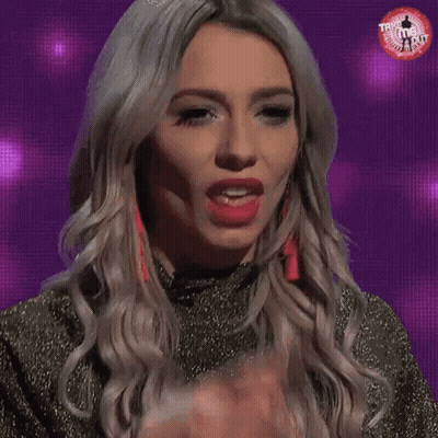 A Blonde Woman Is Licking Her Finger While Wearing Earrings And A Gold Dress . GIF
