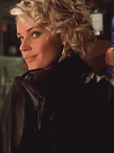 A Blonde Woman Wearing A Leather Jacket And A Blue Top Smiles GIF