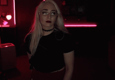 A Blonde Woman Wearing Glasses And A Choker Holds Her Finger To Her Lips GIF