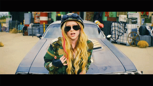 A Blonde Woman Wearing Sunglasses And A Helmet Stands In Front Of A Car GIF