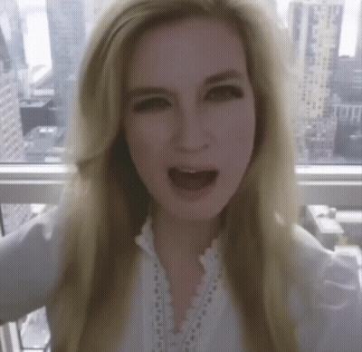 A Blonde Woman With Her Mouth Open Stands In Front Of A Window GIF