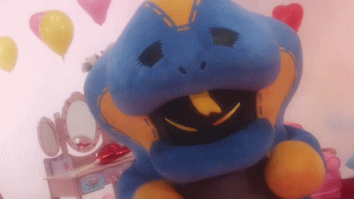 A Blue And Yellow Stuffed Animal Is Sitting In Front Of A Vanity With Balloons In The Background GIF