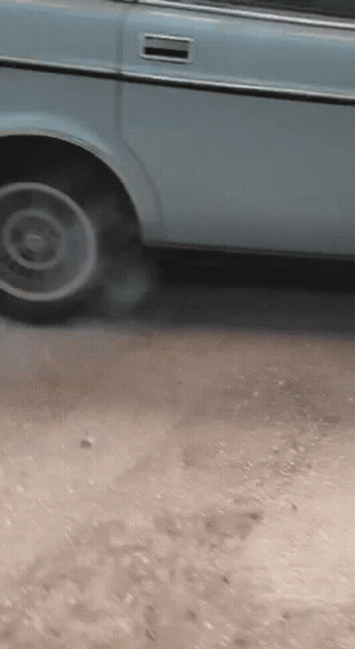 A Blue Car Is Parked On A Dirt Road And Smoke Is Coming Out Of The Tire GIF