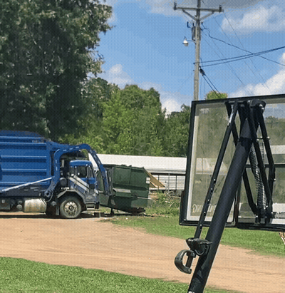 A Blue Garbage Truck Is Parked On A Dirt Road Next To A Sign That Says Exit GIF