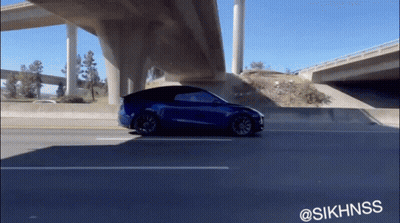 A Blue Tesla Model Y Is Driving Under A Highway Bridge GIF