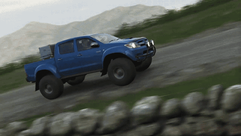 A Blue Truck Driving Down A Dirt Road With Mountains In The Background GIF