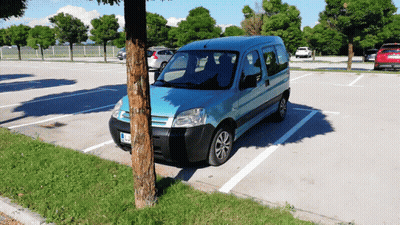 A Blue Van Is Parked In A Parking Lot With A Tree In The Foreground GIF