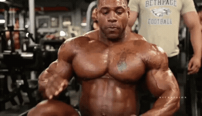 A Bodybuilder Is Flexing His Muscles In A Gym While Wearing A Football Shirt . GIF
