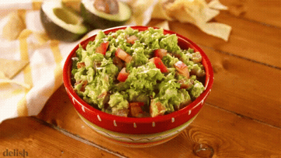 A Bowl Of Guacamole Sits On A Wooden Table Next To A Pile Of Tortilla Chips GIF
