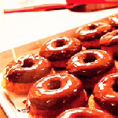 A Box Of Chocolate Covered Donuts With A Red Spoon In The Background GIF
