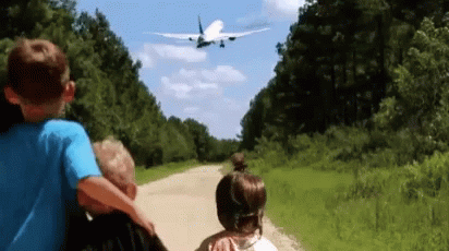A Boy And A Girl Are Looking At An Airplane Flying Over Them GIF
