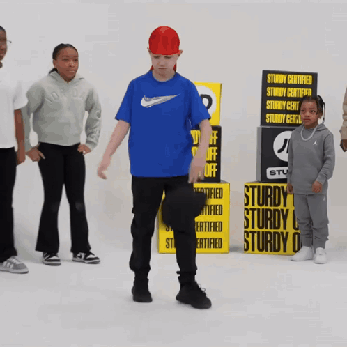 A Boy In A Blue Nike Shirt Stands In Front Of A Stack Of Sturdy Certified Boxes GIF