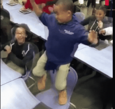 A Boy In A Blue Shirt Is Jumping In The Air While Sitting On A Chair In A Classroom . GIF