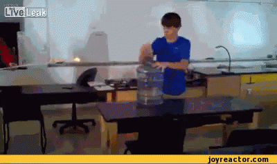 A Boy In A Blue Shirt Is Standing In Front Of A Table With A Bucket Of Water On It .. GIF