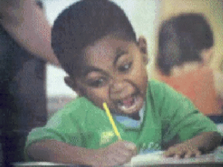 A Boy In A Green Shirt Is Writing On A Piece Of Paper With A Pencil In His Mouth . GIF