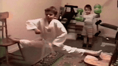 A Boy In A Karate Uniform Is Kicking A Chair While Another Boy Holds A Boxing Glove . GIF