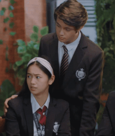 A Boy In A Suit And Tie Stands Next To A Girl In A School Uniform With A Lanyard That Says Homemaker On It GIF