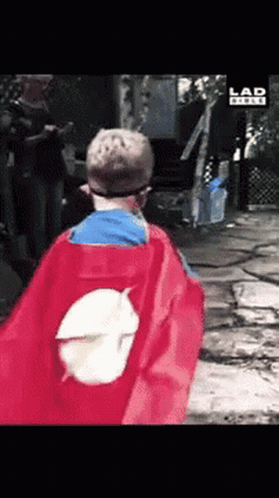 A Boy In A Superhero Cape Is Walking In Front Of A Sign That Says Lad GIF