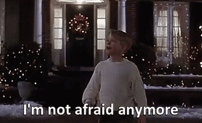 A Boy In A White Sweater Is Standing In Front Of A House With Christmas Lights On It . GIF