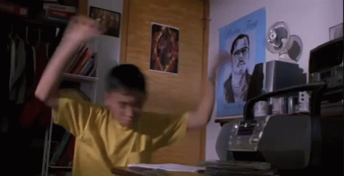 A Boy In A Yellow Shirt Is Sitting At A Desk With His Arms In The Air In A Room . GIF