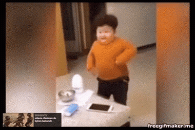 a boy in an orange sweater is standing in front of a table with a cell phone on it