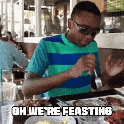 A Boy Is Sitting At A Table With A Plate Of Food And The Words Oh We 'Re Feasting Written Above Him GIF