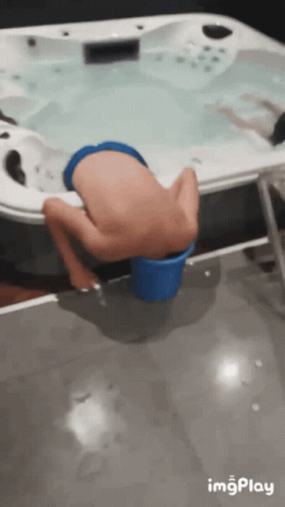 A Boy Is Sitting In A Blue Bucket Near A Hot Tub GIF