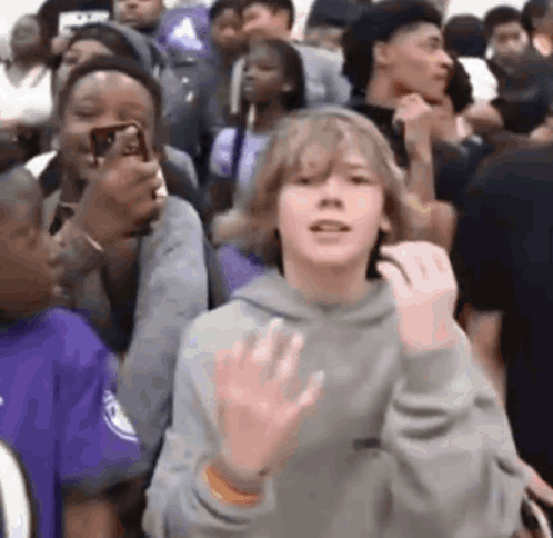 A Boy Is Standing In A Crowd Of People Waving His Hand . GIF