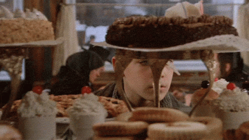 A Boy Is Standing In Front Of A Table Full Of Desserts Including A Chocolate Cake GIF