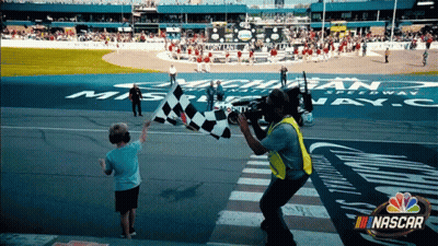 A Boy Waves A Checkered Flag In Front Of A Nascar Advertisement GIF