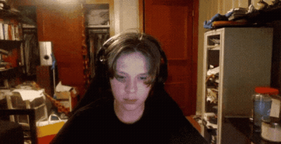 A Boy Wearing Headphones Looks At The Camera In A Cluttered Room GIF