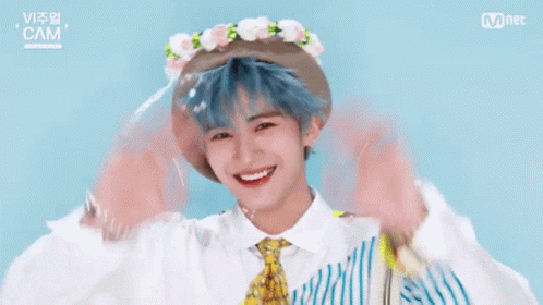 A Boy With Blue Hair And A Flower Crown On His Head Is Waving His Hands . GIF