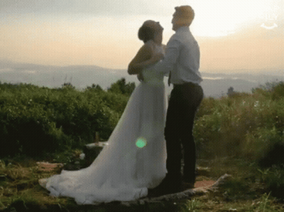 A Bride And Groom Are Dancing In A Field At Sunset . The Bride Is Wearing A Wedding Dress . GIF