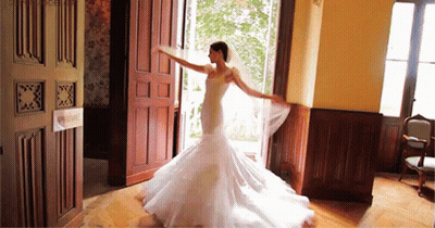A Bride In A Wedding Dress Is Dancing In A Room With A Door Open . GIF