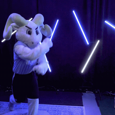 A Bunny Mascot Is Holding Two Lightsabers In Front Of A Blue Curtain GIF
