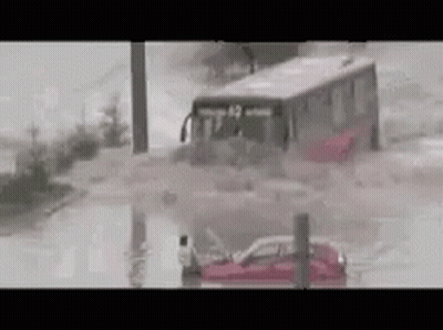 A Bus Is Driving Through A Flooded Street With A Car In The Water . GIF