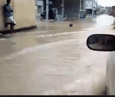 A Car Is Driving Down A Flooded Street With A Man Leaning Against A Wall In The Background GIF