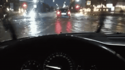 A Car Is Driving Down A Wet Street At Night With A Steering Wheel In The Foreground . GIF
