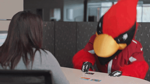 A Cardinal Mascot Is Playing Cards With A Woman At A Table GIF