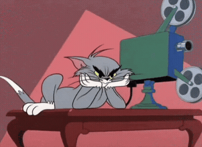 A Cartoon Cat Is Sitting At A Table Watching A Movie On A Film Camera . GIF