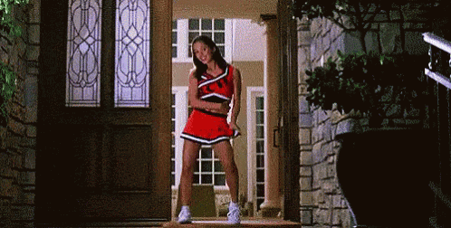 A Cheerleader Is Standing In Front Of A Door Wearing A Red Skirt . GIF