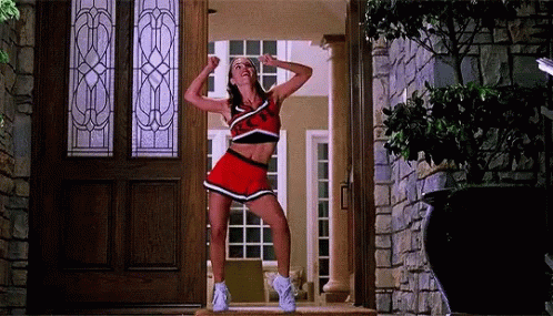 A Cheerleader Is Standing In Front Of A Door With Her Arms In The Air . GIF
