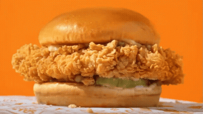 A Close Up Of A Fried Chicken Sandwich On A Bun On A Table GIF