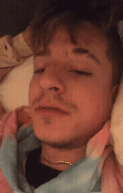 A Close Up Of A Man Laying In Bed With His Eyes Closed And A Tie Dye Shirt On . GIF