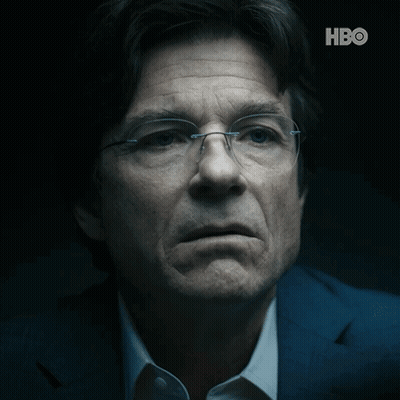 A Close Up Of A Man Wearing Glasses And A Suit With The Hbo Logo Behind Him GIF