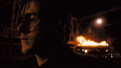 A Close Up Of A Person 'S Face With A Burning Vehicle In The Background GIF