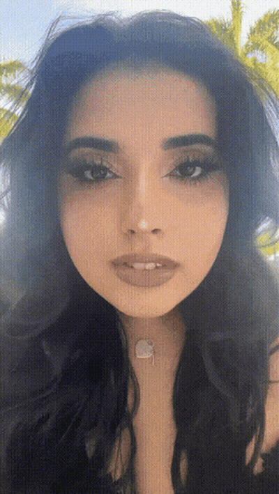 A Close Up Of A Woman 'S Face With A Necklace Around Her Neck And A Palm Tree In The Background GIF