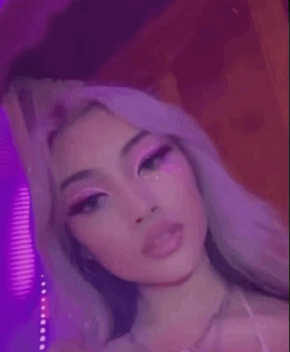 A Close Up Of A Woman 'S Face With Pink Makeup And Purple Lights Behind Her . GIF